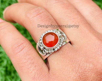 May include: A silver ring with an orange carnelian gemstone set in a decorative bezel. The ring has a scroll-like design and is set on a finger.