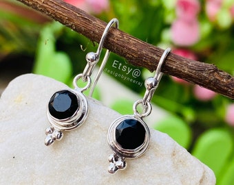以下が含まれることがあります： 黒い宝石のアクセントが付いたシルバーのドロップイヤリングのペア。イヤリングは、シルバーのベゼルにセットされた小さな丸い宝石でシンプルなデザインになっています。イヤリングはシルバーのフックから吊り下げられています。