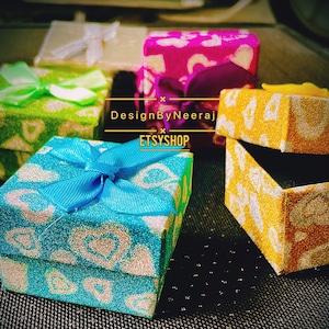 Puede incluir: Cuatro peque&ntilde;as cajas de regalo con purpurina y dise&ntilde;os estampados. Las cajas son de varios colores, incluyendo verde, azul, rosa y amarillo. Las cajas tienen lazos y est&aacute;n dispuestas sobre una superficie negra. El texto "DesignByNeeraj ETSYSHOP" es visible en la imagen.