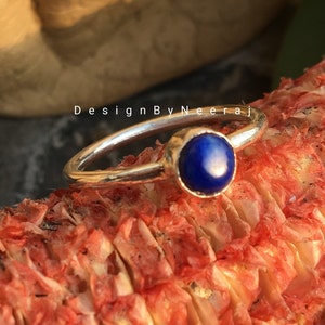 May include: A silver ring with a blue gemstone set in a bezel setting. The ring is resting on a red and white textured surface. "DesignByNeeraj" is written on the surface.