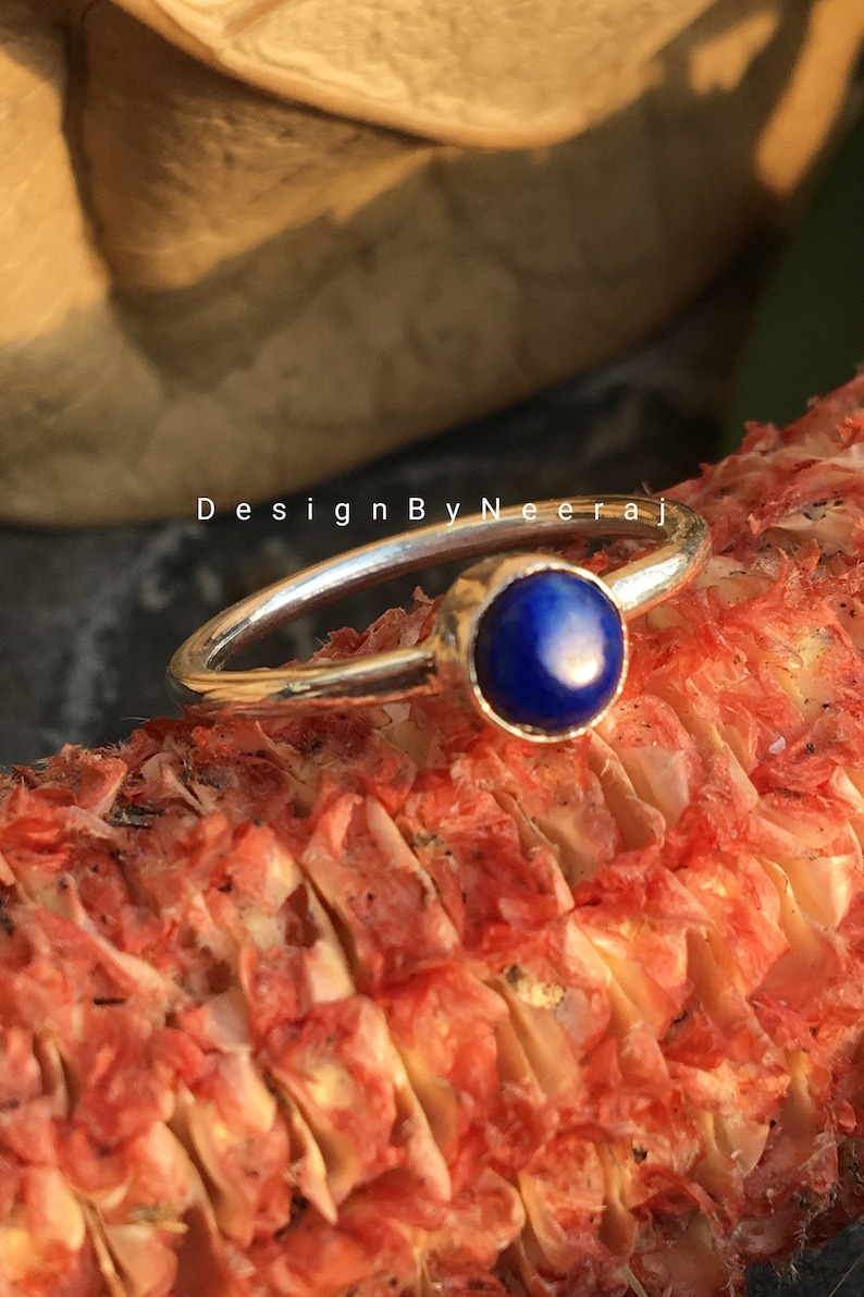 May include: A silver ring with a blue gemstone set in a bezel setting. The ring is resting on a red and white textured surface. "DesignByNeeraj" is written on the surface.