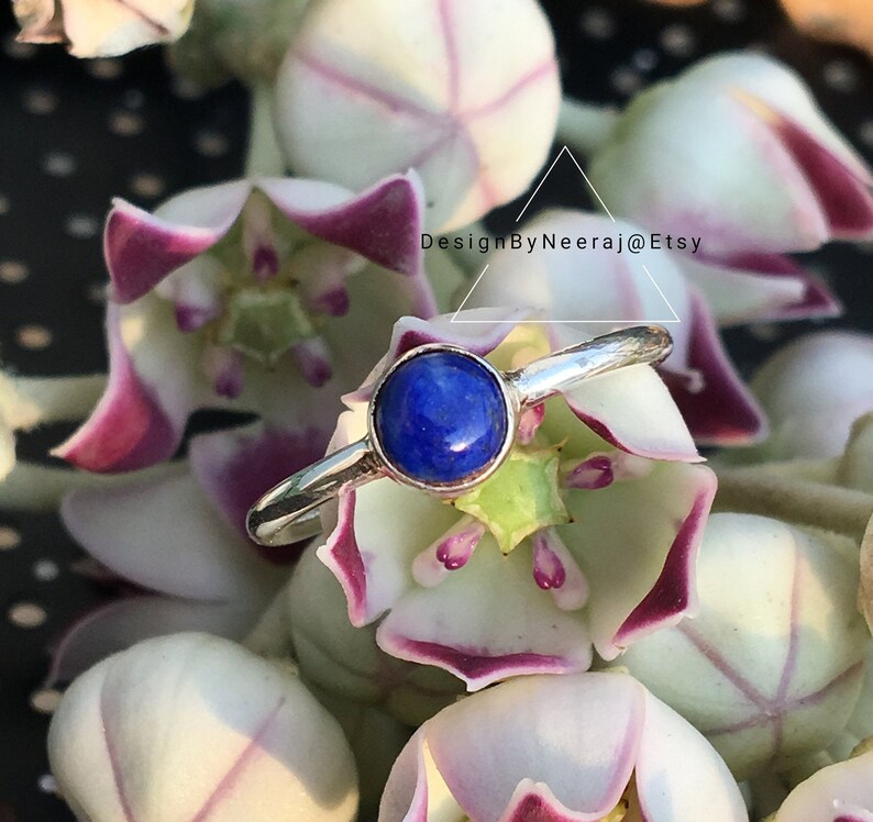 May include: A silver ring with a blue lapis lazuli gemstone. The ring is set against a background of white and purple flowers.
