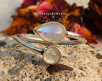 May include: A silver ring with two moonstone gems. The ring is designed to be worn on the finger with the gems facing up. The ring is a delicate and elegant piece of jewelry.