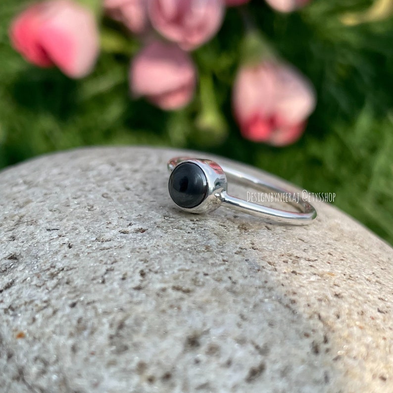 Puede incluir: Un anillo de plata con una piedra preciosa negra engastada en un engaste de bisel. El anillo est&aacute; sobre una roca gris con flores rosas en el fondo.