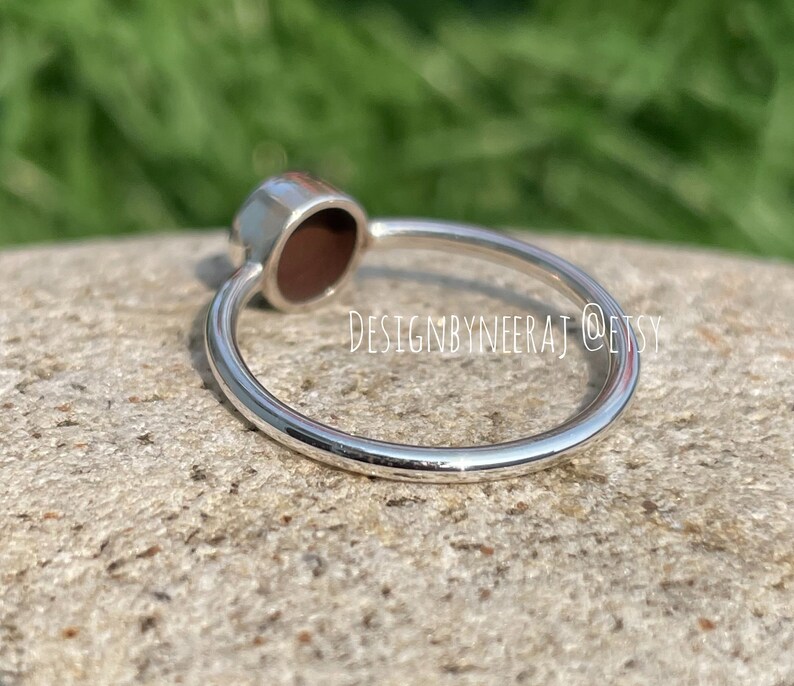 Puede incluir: Un anillo de plata con una piedra ovalada de color rojo oscuro. El anillo est&aacute; sobre una roca gris.