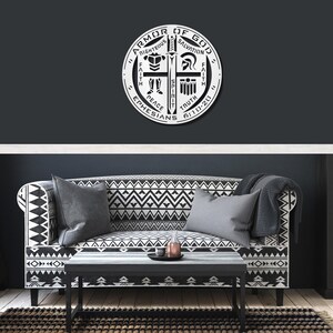 May include: A circular, white metal wall art piece with the words "Armor of God" and other religious terms. The artwork is mounted on a dark gray wall above a patterned sofa and coffee table.