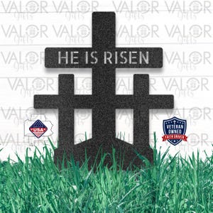 May include: A black metal cross sculpture with the text "HE IS RISEN". The sculpture features three crosses, with the central cross being the largest. The sculpture is set against a white background with green grass in the foreground.