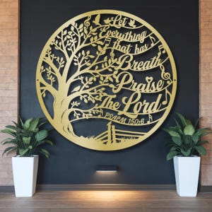 May include: A gold-colored metal wall art piece in a circular design, featuring a tree with musical notes and the words "Let Everything That Has Breath Praise The Lord" and "Psalm 150:6". The art is mounted on a dark wall.