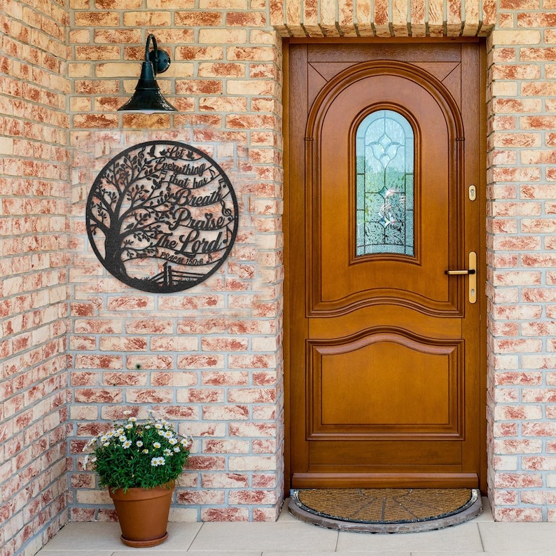 May include: A black metal wall art piece with a tree design and the words "Let everything that has breath praise the Lord" in a circular frame. The art is mounted on a brick wall near a wooden door.