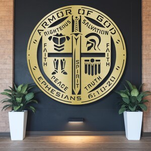 May include: A gold-colored metal wall art piece featuring the words "Armor of God" and other religious text. The design includes a sword, cross, and various symbols. The artwork is mounted on a dark wall with plants in white planters on either side.