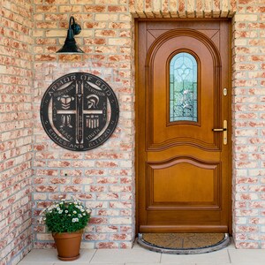 May include: A round, black metal wall hanging with the text "ARMOR OF GOD" and other religious phrases. The sign is mounted on a brick wall next to a wooden door with a glass window. A potted plant sits below the sign.