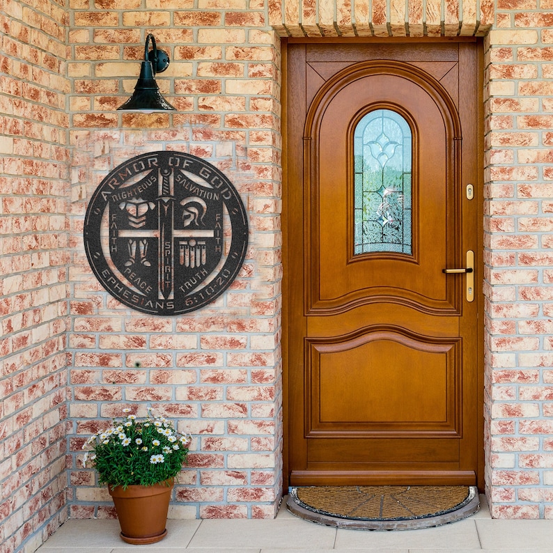 May include: A round, black metal wall hanging with the text "ARMOR OF GOD" and other religious phrases. The sign is mounted on a brick wall next to a wooden door with a glass window. A potted plant sits below the sign.