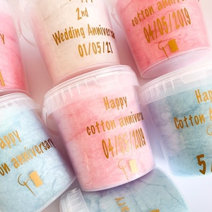 Pu&ograve; includere: Una collezione di sei contenitori di plastica trasparente riempiti di zucchero filato rosa, blu e bianco. Ogni contenitore ha un'etichetta dorata con una data e un messaggio diversi, come "Happy cotton anniversary 04/05/2019" e "2nd Wedding Anniversary 01/05/21".