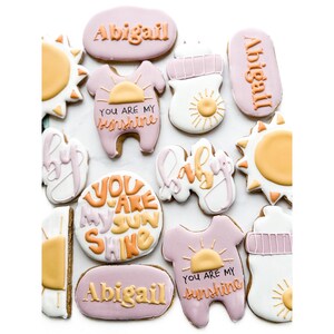 Peut inclure: Assortiment de biscuits au sucre décorés de différentes formes et couleurs. Les biscuits incluent le nom "Abigail", des biberons, des soleils et des bodies avec le texte "You are my sunshine". Les biscuits sont décorés avec du glaçage rose, orange, jaune et blanc.