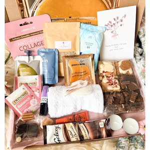 Op de afbeelding: Een geschenkdoos met diverse artikelen, waaronder een roze anti-aging collageen gezichtsmasker, een gouden oogmasker, een witte handdoek, een zakje Galaxy instant warme chocolademelk, Twinings theezakjes, een verjaardagskaart en diverse brownies.