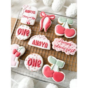 Puede incluir: Una colección de galletas de azúcar decoradas con temática de cerezas. Las galletas tienen varias formas, incluyendo cerezas, lazos y rectángulos, y presentan el nombre "ANAYA" y la palabra "ONE" en glaseado rojo. Las galletas están dispuestas sobre una rejilla.
