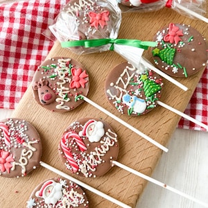 May include: Chocolate Christmas lollipops with festive decorations, including reindeer, snowmen, Christmas trees, and Santa Claus designs. Some lollipops are wrapped in clear cellophane and tied with ribbons. Sprinkles and candy canes add to the holiday theme.
