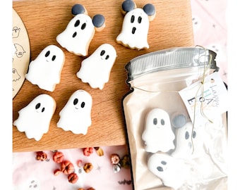 May include: A collection of Halloween-themed ghost cookies. Some cookies have black mouse ears. The cookies are white with black eyes and mouths. Some cookies are on a wooden board, and others are in a clear bag with a tag that says "Happy Halloween".
