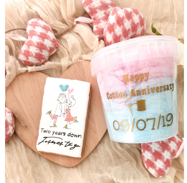 Pu&ograve; includere: Un contenitore di zucchero filato rosa e blu con il testo "Happy Cotton Anniversary 09/07/19". In primo piano, un biscotto decorato con una coppia che tiene dei palloncini e il testo "Two years down forever to go".