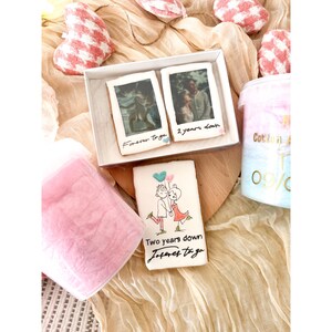 Pu&ograve; includere: Un set regalo per una coppia che celebra il suo anniversario. Il set include due biscotti decorati, uno con una foto della coppia e il testo "Forever together" e l'altro con una foto della coppia e il testo "Two years down". Il set include anche un contenitore di zucchero filato rosa.