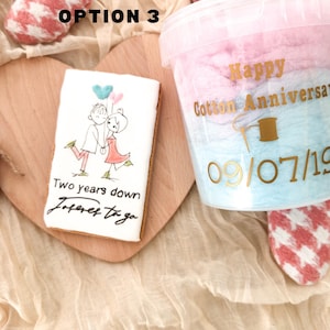 Pu&ograve; includere: Un biscotto bianco con un'illustrazione disegnata a mano di una coppia che tiene dei palloncini. Il testo sul biscotto recita "Two years down, forever to go". Nell'immagine &egrave; presente anche un contenitore di zucchero filato rosa e blu con il testo "Happy Cotton Anniversary 09/07/19".