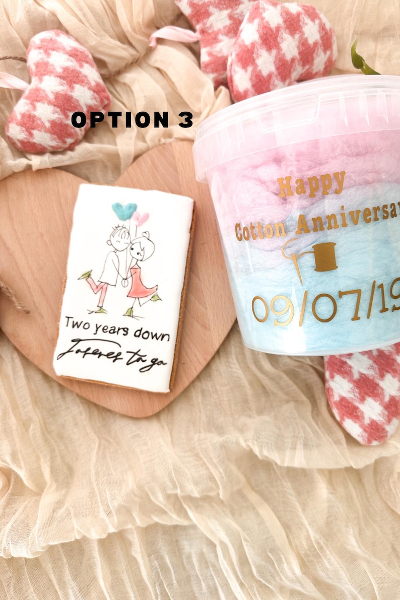 Pu&ograve; includere: Un biscotto bianco con un'illustrazione disegnata a mano di una coppia che tiene dei palloncini. Il testo sul biscotto recita "Two years down, forever to go". Nell'immagine &egrave; presente anche un contenitore di zucchero filato rosa e blu con il testo "Happy Cotton Anniversary 09/07/19".
