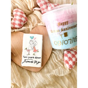 Pu&ograve; includere: Un biscotto decorato con una coppia che tiene palloncini e il testo "Two years down forever to go". Un contenitore di zucchero filato rosa e blu con il testo "Happy Cotton Anniversary 09/07/19".