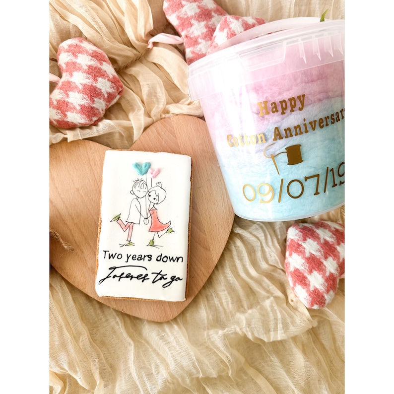 Pu&ograve; includere: Un biscotto decorato con una coppia che tiene palloncini e il testo "Two years down forever to go". Un contenitore di zucchero filato rosa e blu con il testo "Happy Cotton Anniversary 09/07/19".