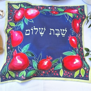 May include: A blue fabric challah cover with a yellow border. The cover is decorated with pomegranates and green leaves. The Hebrew text "Shabbat Shalom" is written in white on the cover.