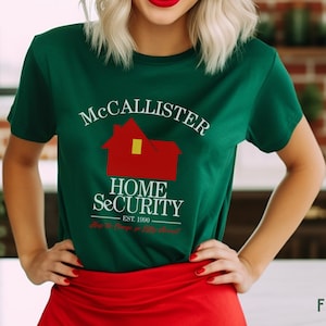 May include: A forest green t-shirt with the text "McCallister Home Security" and a red house graphic. The shirt also includes the text "Est. 1990" and a tagline. The model is wearing a red skirt.