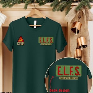 May include: Dark green t-shirt with the text "E.L.F.S. ELVES WITH ATTITUDE" on the front and back. The front also features a red and yellow triangle with the word "NOPO". The shirt is displayed on a wooden hanger.