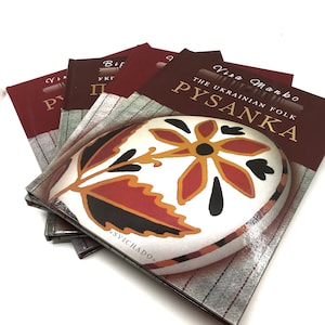 May include: A stack of five books with red covers and gold lettering. The top book features a colorful illustration of a pysanka, a traditional Ukrainian Easter egg, with a red, yellow, and black design. The title of the book is "Vira Manko The Ukrainian Folk Pysanka".