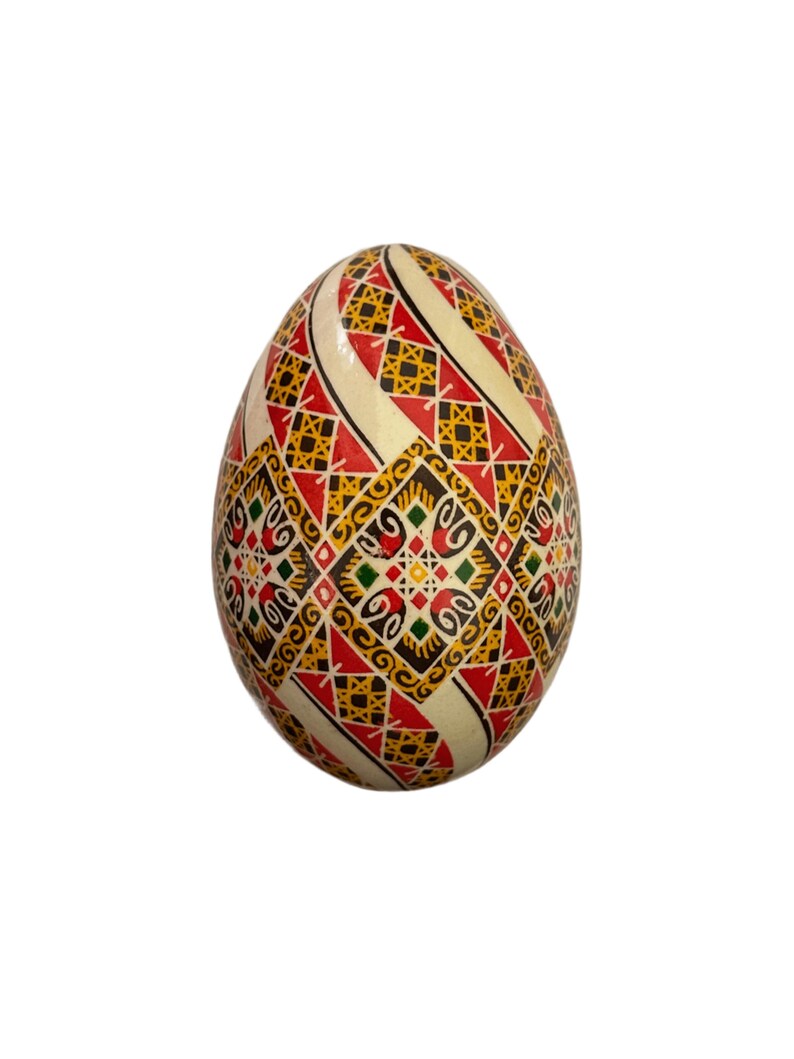 May include: A white egg decorated with a geometric pattern in red, yellow, black, and green. The pattern is intricate and includes squares, diamonds, and swirls.