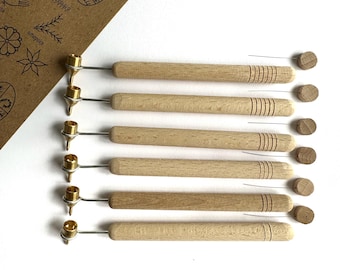 May include: Six wooden-handled tools with metal tips, possibly for crafting or art, are arranged on a white surface. A brown paper with illustrations is in the background. The tools have a natural wood finish. The paper has illustrations.