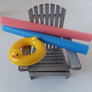 May include: A miniature gray wooden Adirondack chair with a pink and blue pool noodle and a yellow duck-shaped inflatable ring. The pool floats are resting on the chair, suggesting a summer or beach theme.