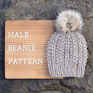 May include: A gray knitted beanie with a fluffy pom pom on top. The beanie has a cable knit pattern and the words "HALE BEANIE PATTERN" are on a wooden sign behind it.