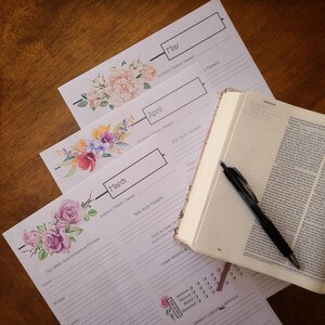 May include: Three floral-themed printable monthly planner pages with sections for Scripture Memory Passage, Bible Study Passages, Truth to Dwell On, Prayer Requests, and Weekend Gratitude & Reflection. The pages are stacked on top of each other, with the month of May on top, followed by April, and then March. A closed Bible and a black pen are lying on top of the pages.