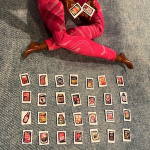 May include: A doll's legs in pink patterned pants holding two small food item cards. Twenty-four miniature food product cards are scattered on a denim surface, featuring various canned goods and packaged items.