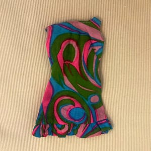 May include: A sleeveless doll dress featuring a vibrant, swirling abstract pattern in shades of bright pink, forest green, and sky blue. The garment has a fitted bodice, a flared skirt, and a small ruffled hemline along the bottom edge.