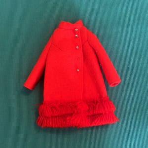 May include: A bright red coat featuring a high collar, long sleeves, and a front button closure with small silver-toned buttons. The hem of the coat is finished with a thick, textured fringe detail that extends across the entire bottom edge.
