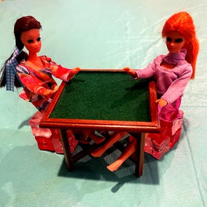 May include: Two dolls are seated at a miniature wooden table with a green felt top. The dolls are dressed in colorful outfits. The table is brown and has a classic design.