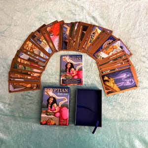 May include: An open deck of Egyptian Oracle Cards fanned out, with two boxes and a blue box with a ribbon. The card backs are brown, and the fronts feature colorful illustrations. The boxes read "Egyptian Oracle Cards."