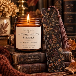 May include: An 8 oz soy blend candle in an amber glass jar with a white label reading Tale of Wicks Autumn Nights & Books, accompanied by a rectangular bookmark featuring a dark floral pattern and a brown tassel, resting on vintage books.