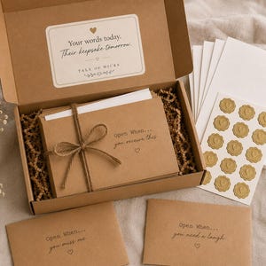 May include: A brown cardboard gift box containing a stack of kraft paper envelopes tied with twine, labeled Open When... you receive this. Includes white envelopes, a sheet of gold heart stickers, and two loose envelopes labeled for specific occasions.