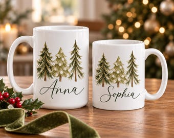 May include: Two white ceramic mugs featuring a watercolor illustration of three pine trees with white accents. The larger mug displays the name Anna in black script, while the smaller mug features the name Sophia in black script.