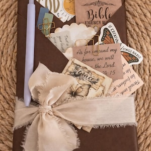 May include: A brown journal with a cream-colored ribbon and a pen. The journal's pocket holds cards with text, including "Bible" and "Grace", along with butterfly illustrations. Gold paper clips and a tag that says "Where God" are also visible.