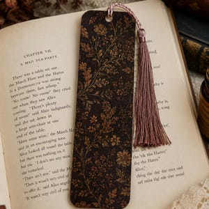 Moody autumn botanical bookmark with dusty mauve tassel resting on an open vintage book, featuring a dark brown floral design with rust, burgundy and golden botanical details in a cozy fall reading scene.