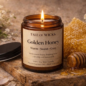Puede incluir: Una vela Golden Honey encendida en un tarro de cristal marrón con tapa negra, junto a un panal y un cucharón de miel de madera. La etiqueta dice "Tale of Wicks" y "Golden Honey".