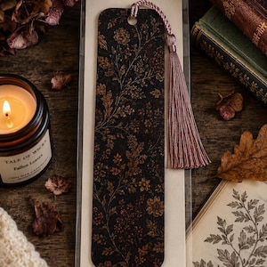 Moody autumn botanical bookmark with dusty mauve tassel displayed in gift-ready packaging beside a glowing fall candle, vintage books, dried flowers and autumn leaves on a rustic wood table.