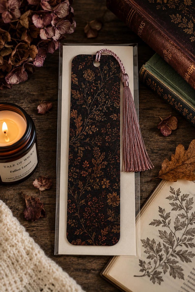 Moody autumn botanical bookmark with dusty mauve tassel displayed in gift-ready packaging beside a glowing fall candle, vintage books, dried flowers and autumn leaves on a rustic wood table.